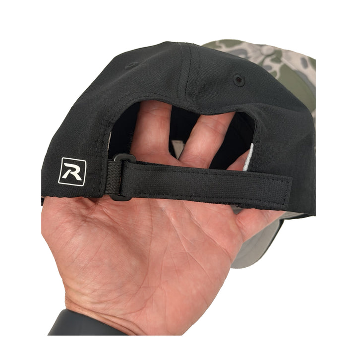 Translation missing: en.Close-up of the back of a black baseball cap being held in a hand, showing the adjustable black strap and a small white embroidered logo patch. The brim edge reveals a camouflage pattern, and the background is solid white
