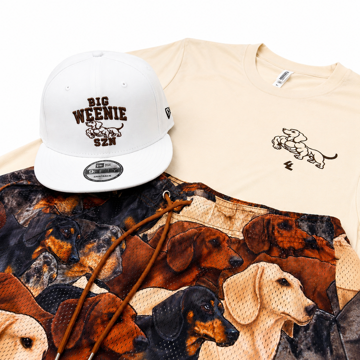 Translation missing: en.A flat-lay product presentation featuring a white New Era baseball cap with 'BIG WEENIE SZN' embroidery, a cream t-shirt with a dachshund graphic, and a pair of shorts with a dachshund print and brown drawstrings, all on a white background