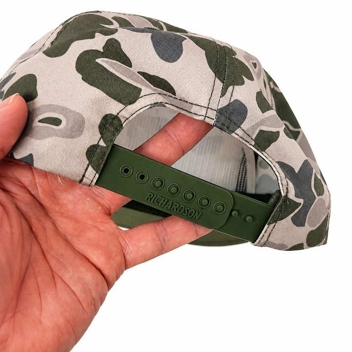 Translation missing: en.A close-up of a camouflage snapback cap being held by a hand, showing the green adjustable strap with 'RICHARDSON' embossed on it, against a plain white background