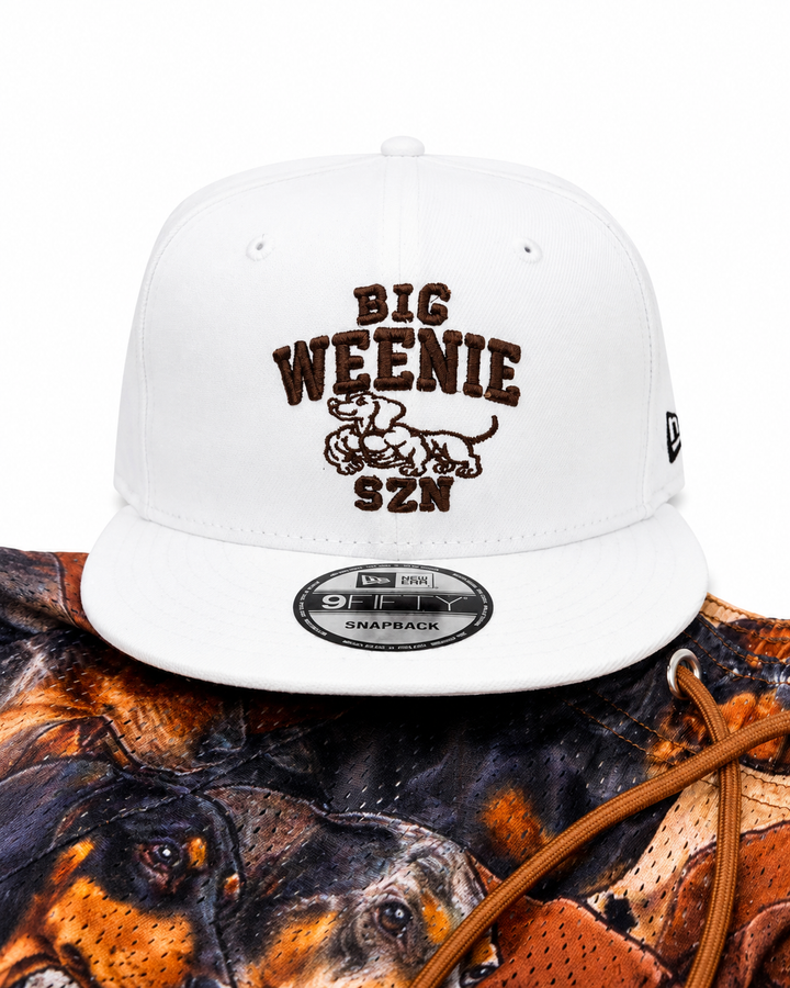 Translation missing: en.A white flat-brim baseball cap with brown embroidered text reading 'BIG WEENIE SZN' and a dachshund graphic on the front. The cap is shown resting on a dog-print shorts with brown drawstrings against a plain white background