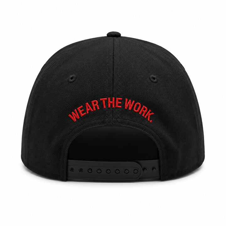 Translation missing: en.Black snapback cap shown from the back, featuring the phrase 'WEAR THE WORK.' embroidered in red arched text across the back panel