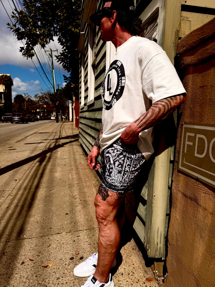 Translation missing: en.A man stands on a city sidewalk wearing a white graphic t-shirt, black and white patterned shorts, white sneakers, and a black cap, leaning against a building