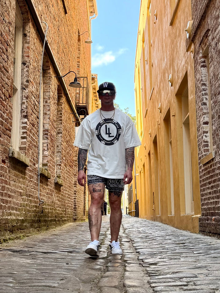 Translation missing: en.A man walking down a narrow cobblestone alleyway between brick buildings, wearing a white t-shirt with a black circular graphic, patterned shorts, and white sneakers