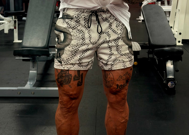 Translation missing: en.A man in a gym taking a mirror selfie, wearing a white t-shirt, a black beanie, and black-and-white snakeskin print shorts. He is holding an orange smartphone