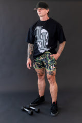 Person wearing a black t-shirt with a logo and camouflage shorts on a dark background
