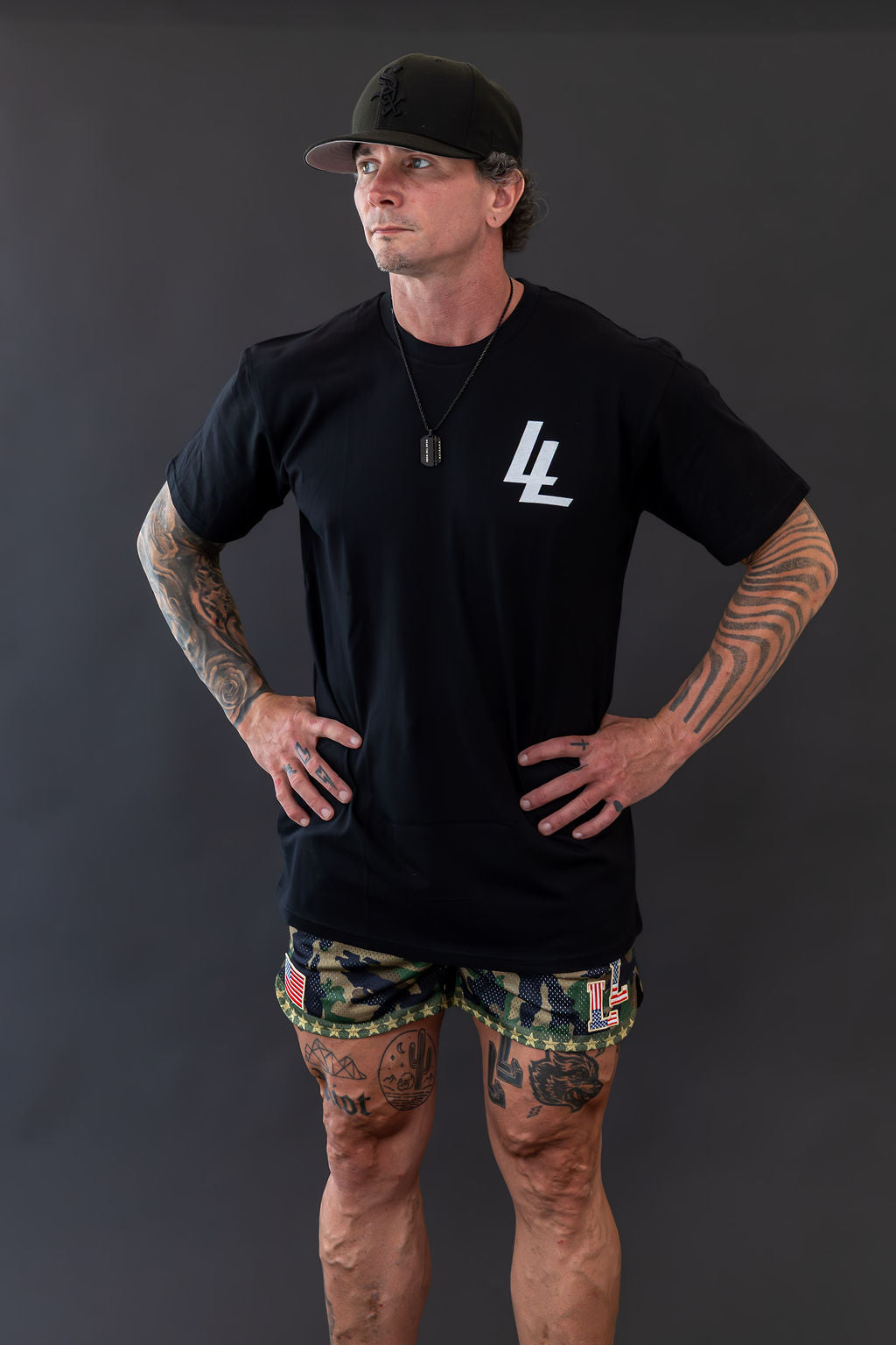 Man wearing a black t-shirt with a logo and patterned shorts on a dark background