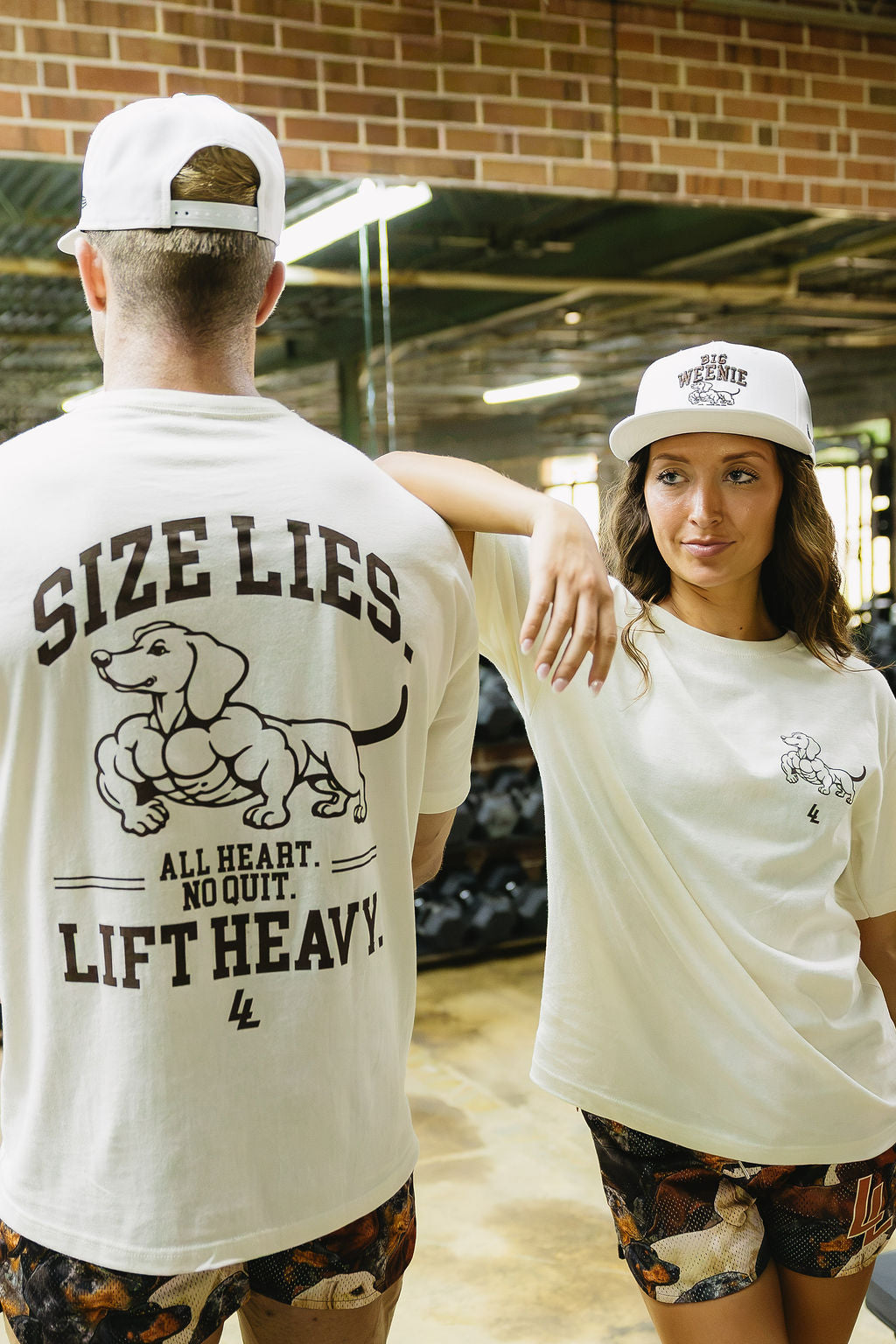 Translation missing: en.Two models wearing a cream graphic t-shirt and a white baseball cap in a gym setting. The model on the left shows the back of the t-shirt with a muscular dachshund graphic and the text 'SIZE LIES