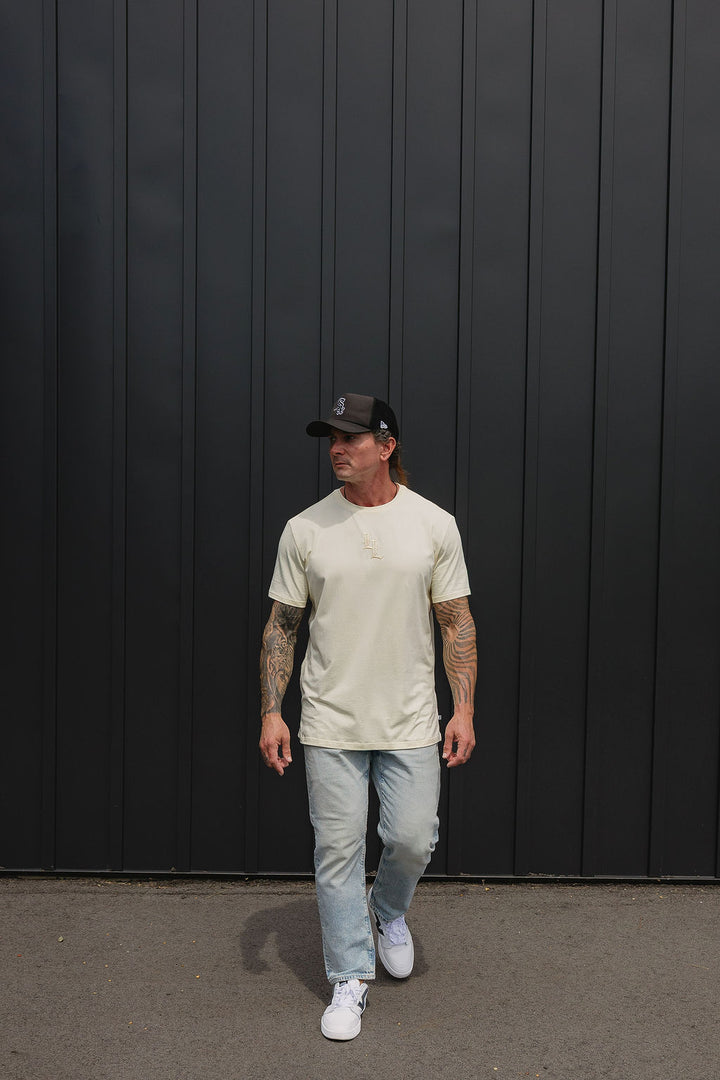 Translation missing: en.A man wearing a light beige crewneck t-shirt, light blue jeans, white sneakers, and a black baseball cap walks in front of a dark gray vertical-paneled wall