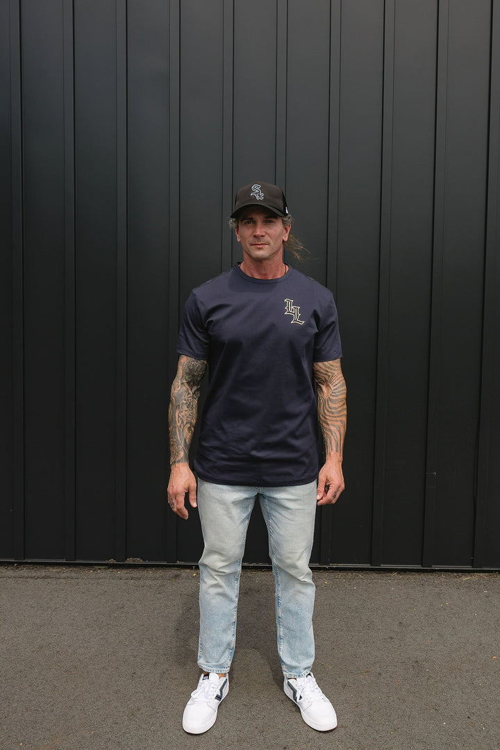 Translation missing: en.A man wearing a navy blue t-shirt with a small white graphic on the left chest, light wash jeans, white sneakers, and a black baseball cap, standing in front of a dark gray vertical-paneled wall