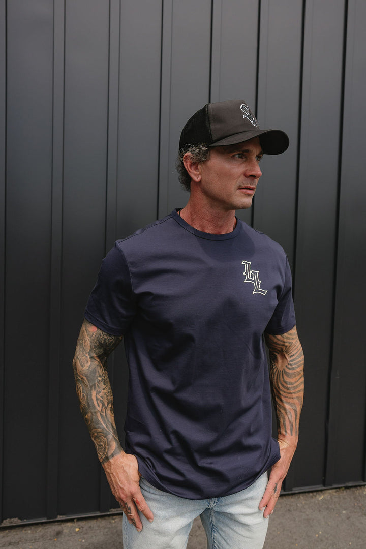 Translation missing: en.A man wearing a navy blue crewneck t-shirt with a white embroidered 'LL' logo on the chest, paired with a black baseball cap and light wash jeans, standing against a dark gray vertical-paneled wall