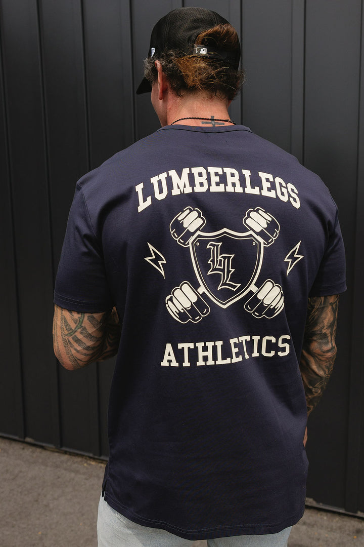 Translation missing: en.A man viewed from behind wearing a navy blue t-shirt with a large white graphic on the back that reads 'LUMBERLEGS ATHLETICS' around a shield logo, standing in front of a dark gray paneled wall
