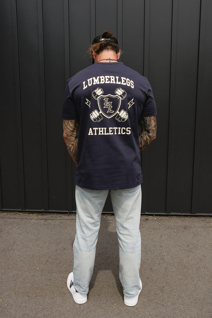 Translation missing: en.A man viewed from behind wearing a navy blue t-shirt with a large white graphic on the back that reads 'LUMBERLEGS ATHLETICS' and light-wash jeans, standing in front of a dark gray vertical-paneled wall