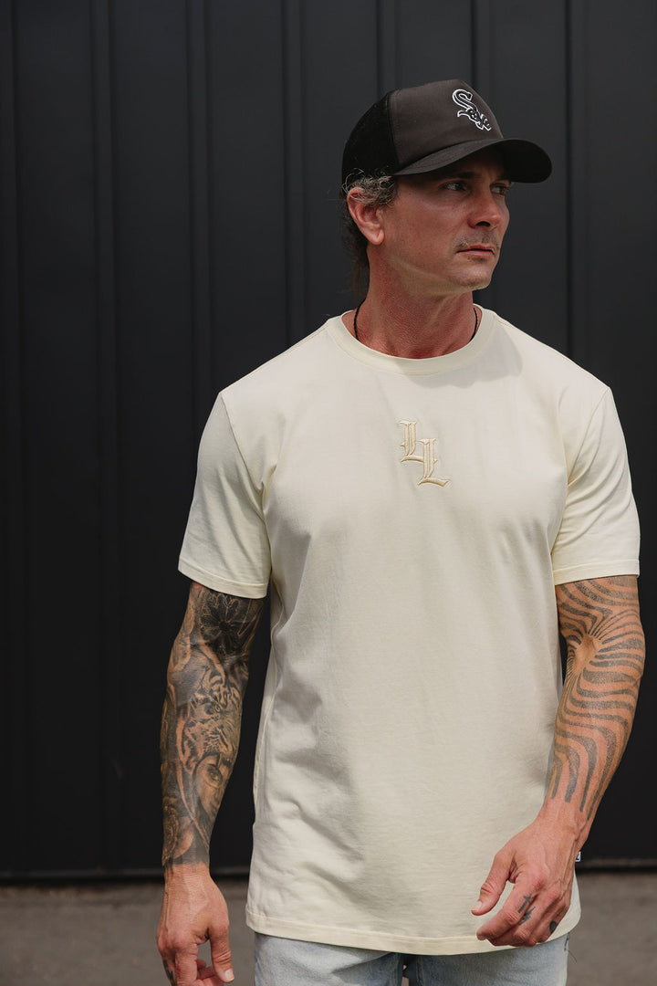 Translation missing: en.A man wearing a cream-colored short-sleeve t-shirt with a small gold embroidered LL  logo on the chest, paired with a black baseball cap and light blue jeans, standing against a black  paneled wall
