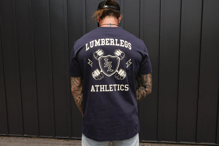 Translation missing: en.A man viewed from behind wearing a navy blue t-shirt with a large white graphic on the back that reads 'LUMBERLEGS ATHLETICS' with a shield and dumbbell design, standing in front of a dark gray vertical slat wall