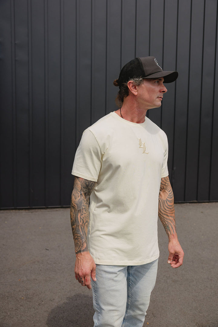 Translation missing: en.A man wearing a cream-colored crewneck t-shirt with an embroidered LL logo  on the center chest, paired with light blue jeans and a black trucker hat, standing in front of a dark gray vertical-paneled wall