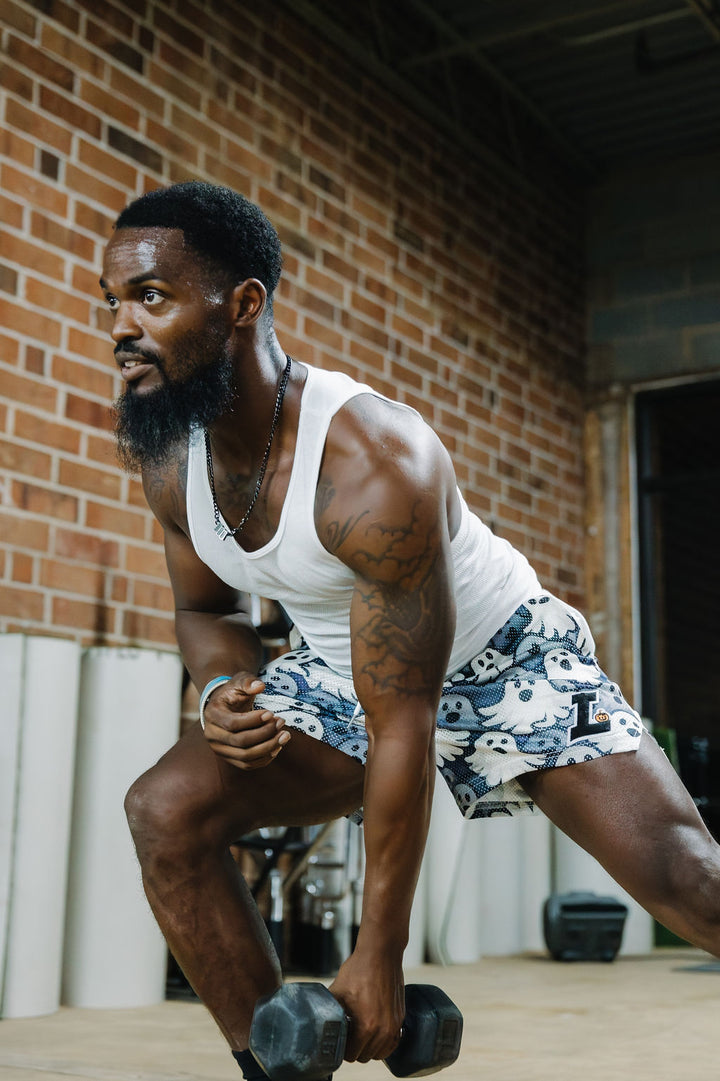 Translation missing: en.A man in a gym wearing a white tank top and grey-and-white all over ghosts patterned shorts, holding a dumbbell while leaning forward in a lunge position against a brick wall background