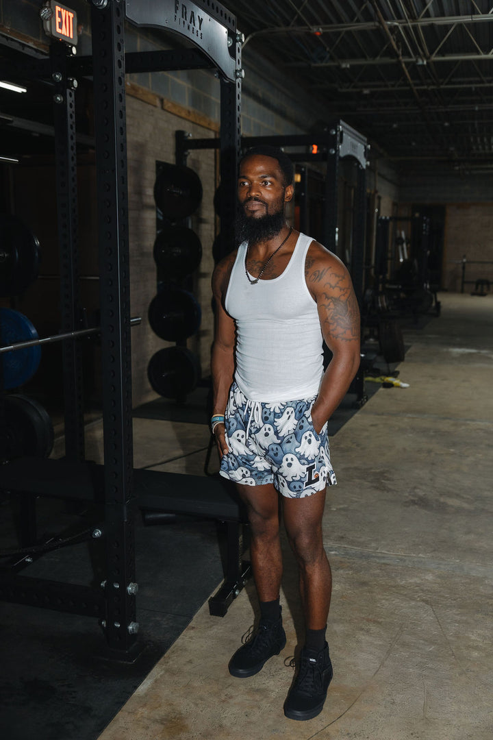 Translation missing: en.A man standing in a gym wearing a white tank top, halloween ghosts patterned athletic shorts, black socks, and black sneakers
