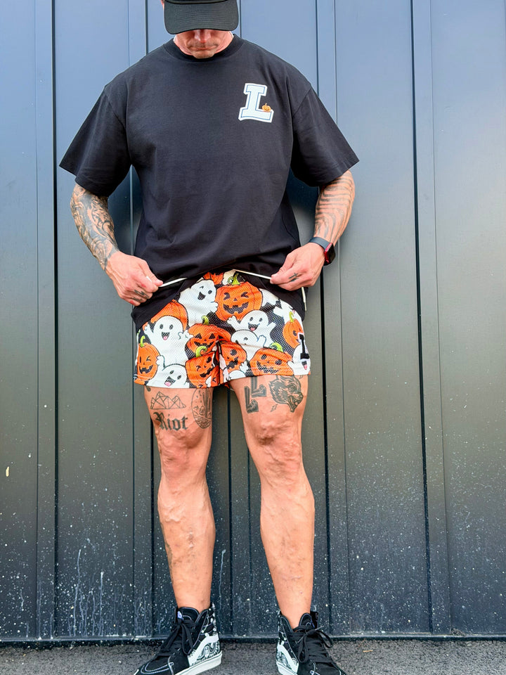 Translation missing: en.A man wearing a black t-shirt and black cap stands against a dark gray metal wall, pulling up black and white patterned athletic shorts with a pumpkin and ghost design