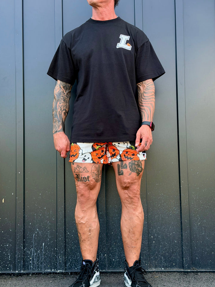 Translation missing: en.A man wearing a black t-shirt with a white and orange 'L' logo on the chest, paired with white athletic shorts featuring a vibrant orange and black pumpkin and ghost halloween pattern, standing against a dark gray metal wall