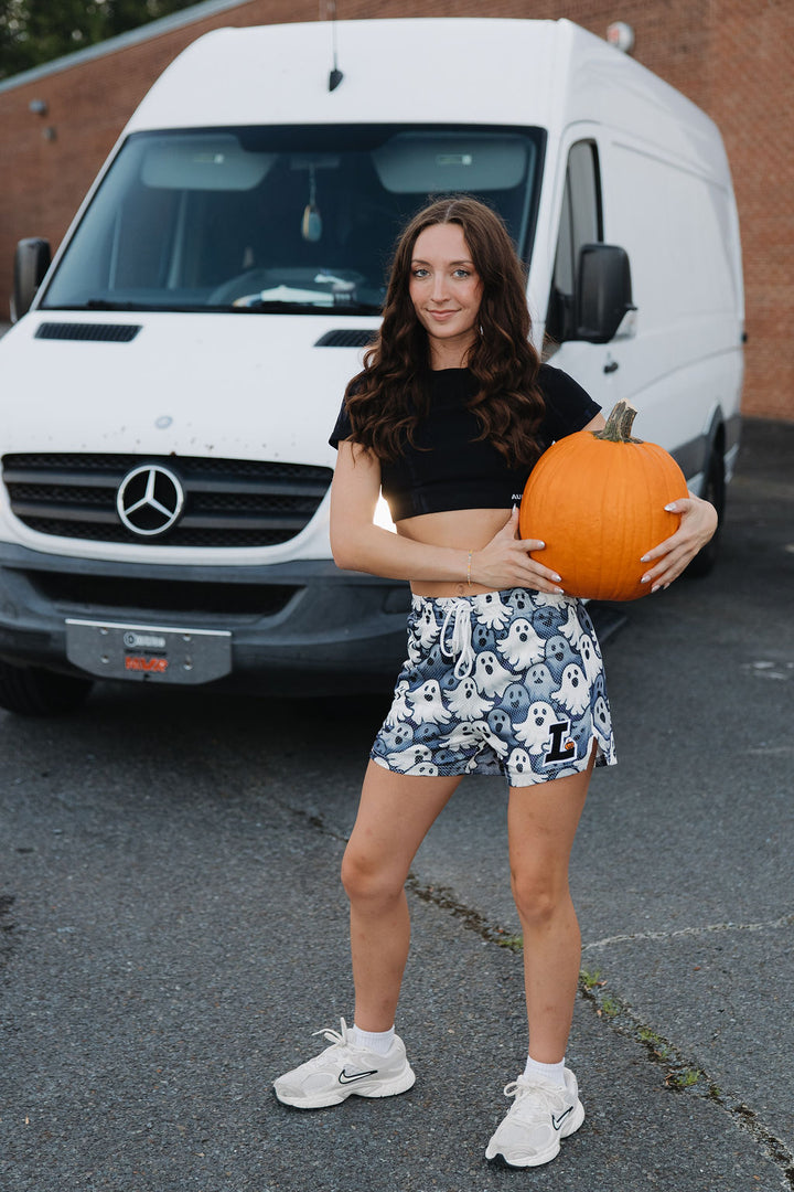 Translation missing: en.A woman stands on asphalt in front of a white Mercedes-Benz Sprinter van, holding a large orange pumpkin. She is wearing a black crop top, grey and white ghosts patterned Halloween shorts, and white sneakers