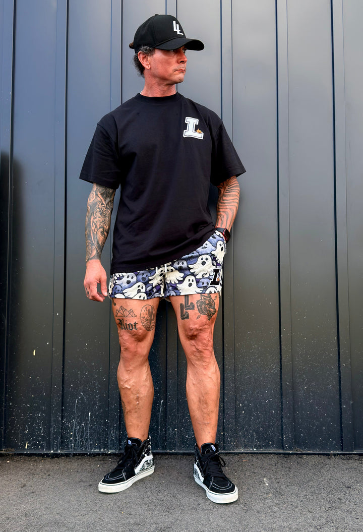 Translation missing: en.A man wearing a black t-shirt with a white L logo with an orange pumpkin graphic, ghosts patterned shorts, a black baseball cap, and black sneakers, standing in front of a dark gray metal wall