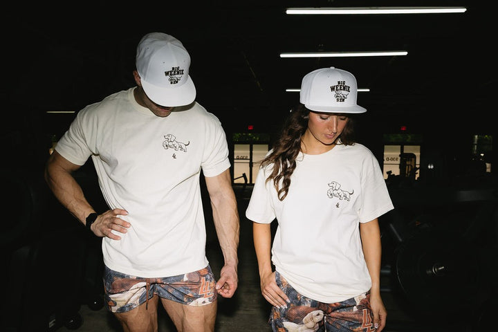 Translation missing: en.A man and a woman standing in a dark gym environment, both wearing matching cream graphic t-shirts, white New Era caps, and dachshund patterned shorts