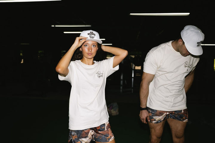 Translation missing: en.Two models wearing matching cream dachshund  t-shirts, white baseball caps, and dachshund pattered mesh gym shorts in a dark indoor gym