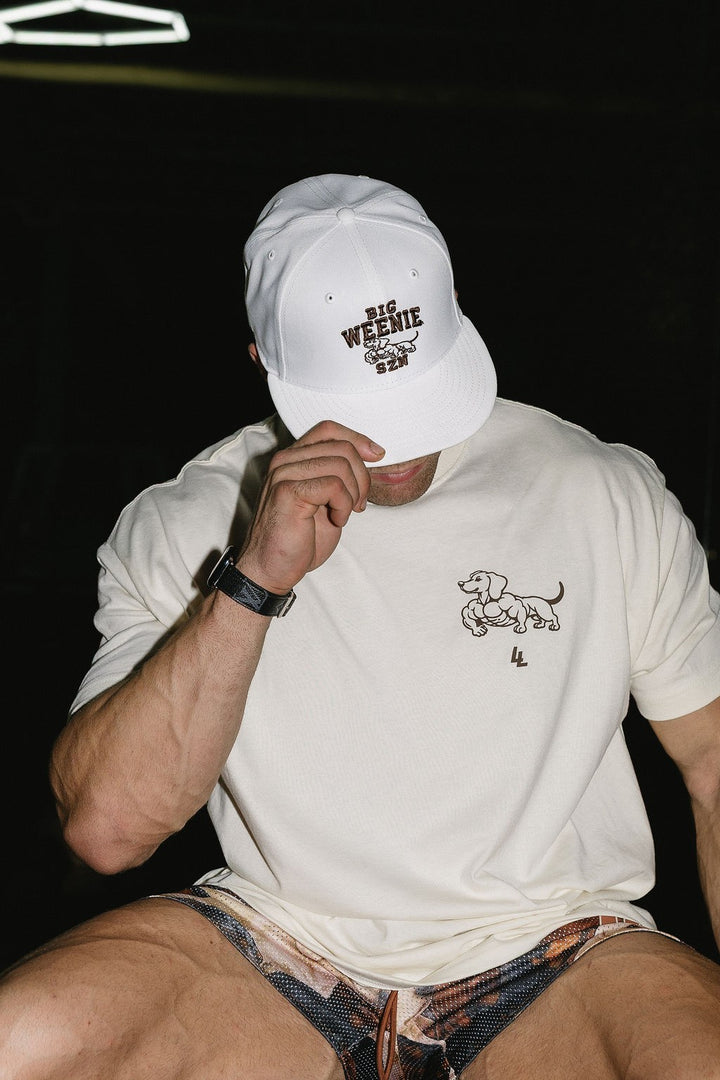 Translation missing: en.A man wearing a cream t-shirt with a swole weenie dog  graphic on the left chest, a white baseball cap with 'BIG WEENIE SZN' text, and dachshund shorts, posed against a dark background