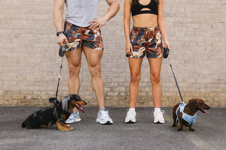 Translation missing: en.A man and a woman standing outdoors in front of a brick wall, both wearing matching multicolor patterned dachshund dog shorts. Two dachshunds are on leashes at their feet, one black and tan and one brown with a blue harness
