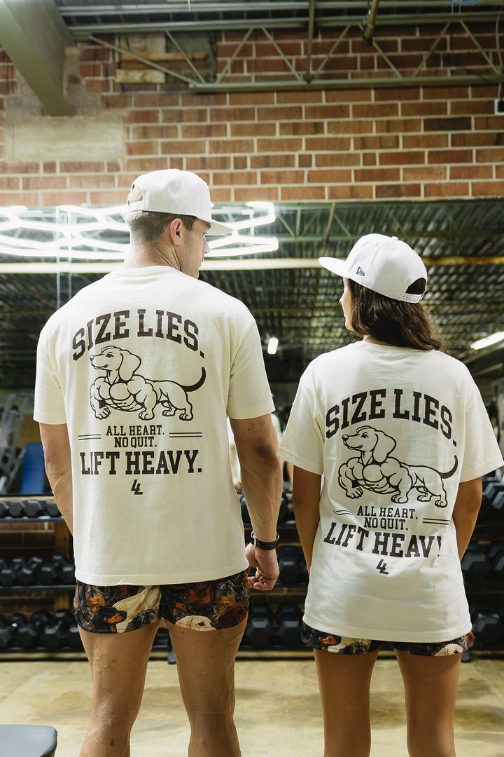 Translation missing: en.Two people standing in a gym, viewed from behind, wearing matching off-white t-shirts with a brown graphic of a dachshund and the text 'SIZE LIES. ALL HEART, NO QUIT, LIFT HEAVY