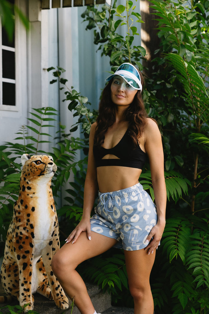 Translation missing: en.A woman posing outdoors in a black sports bra and light blue cheetah. patterned shorts, wearing a blue visor, next to a large cheetah in a garden setting