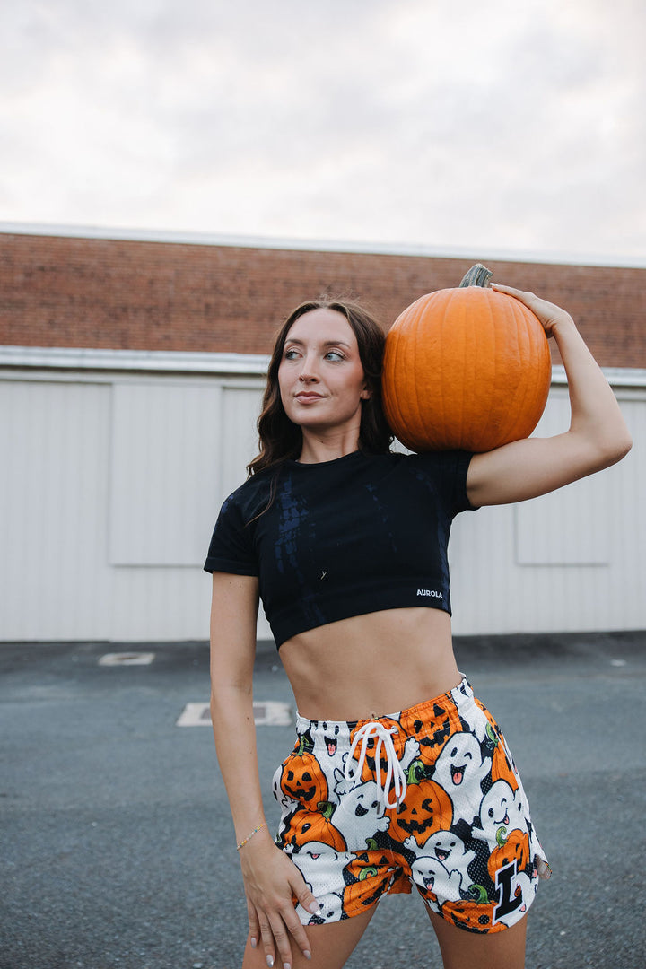 Translation missing: en.A woman posing outdoors in a black crop top and Halloween-themed shorts with a large orange pumpkin on her shoulder, set against a white garage door and brick building background